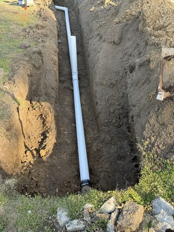 A deep narrow trench with a new white PVC sewer pipe at the bottom