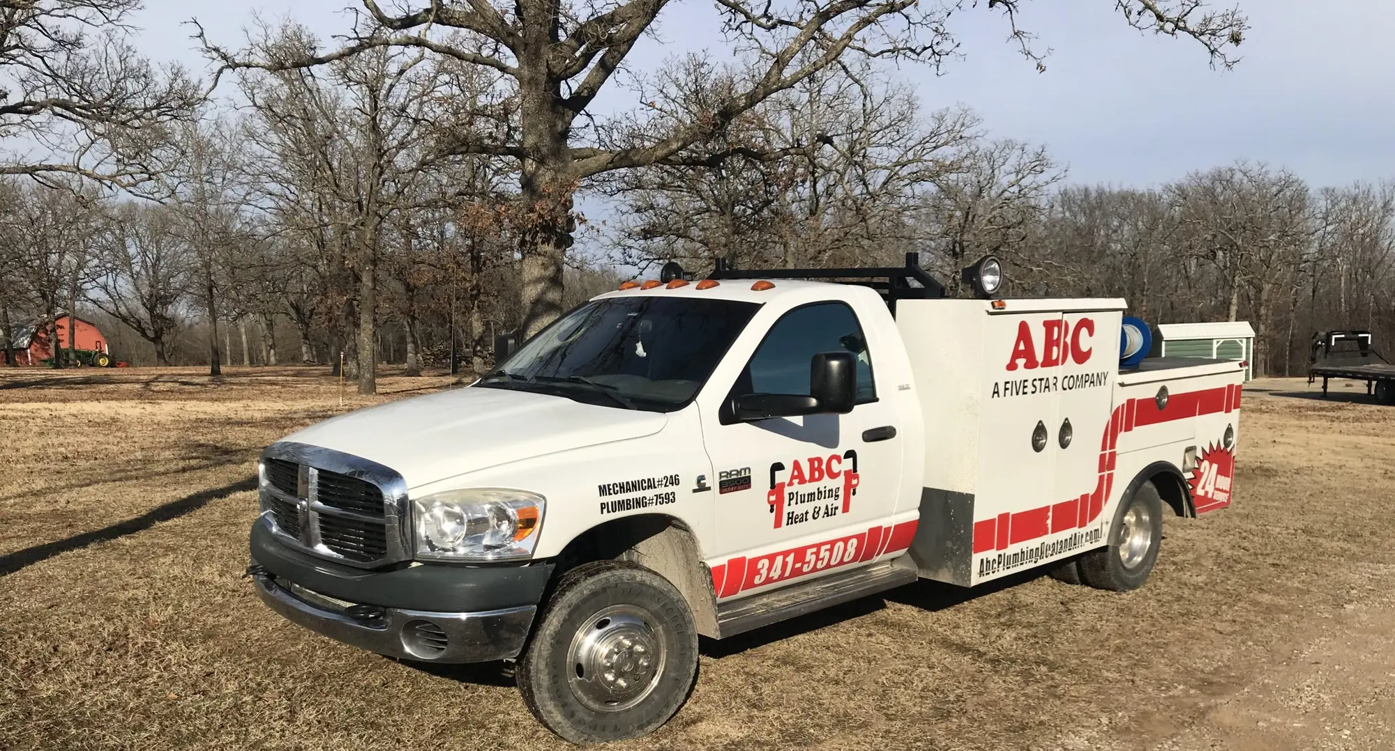 ABC Plumbing Heat and Air's white Dodge service truck with red striping, the license numbers on the door, parked on a gravel lot under bare oak trees